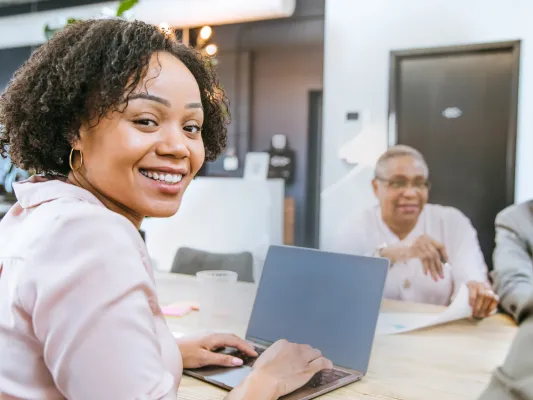 Young woman sitting at a computer, looking at the camera. Coworkers talk in the background.