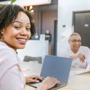 Young woman sitting at a computer, looking at the camera. Coworkers talk in the background.